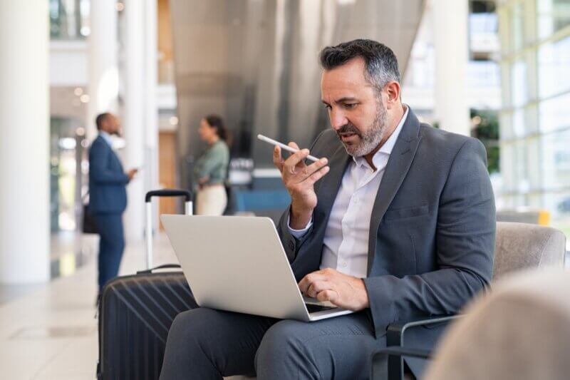 Ein Geschäftsmann sitzt auf einer Couch in einer Flughafen-Lounge, benutzt einen Laptop und sendet eine Sprachnachricht mit seinem Smartphone.